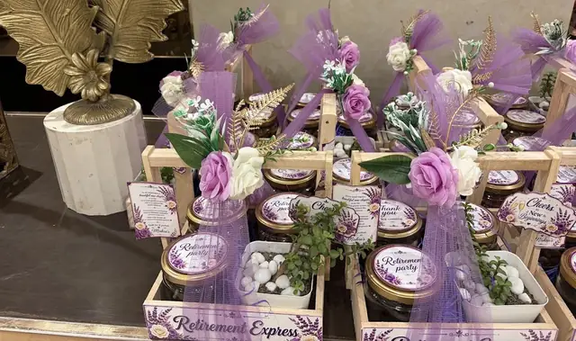 Rows of small wooden crates, each holding a gold-lidded jar, a succulent in white pebbles and a printed tag, dressed with purple tulle and cream and lilac roses.